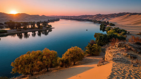 A wide river winds through a desert environment, reflecting the warm hues of a setting sun. The landscape features rolling sand dunes, sparse vegetation, and a clear blue sky transitioning to shades of orange and purple. This image may be suitable for travel, nature, or environmental content.の素材