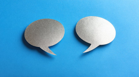 Two speech bubbles are positioned against a bright blue backdrop. The bubbles exhibit a smooth texture with varying shades of gray, suggesting a three-dimensional effect. The composition utilizes simple lighting and focuses directly on the subject, suitable for various editorial and promotional applications.の素材
