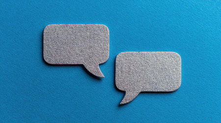 Two silver speech bubbles rest against a vibrant blue backdrop. The shapes present a clean, minimalist design with a textured surface. The composition suggests an overhead view, with even lighting, suitable for a variety of media uses including editorial or commercial design projects. It presents an illustration of communication.の素材