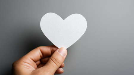 A hand delicately holds a white heart-shaped cutout against a solid gray backdrop. The image features soft lighting and a simple composition, highlighting the heart's form. The scene's neutral tones suggest a versatile use for projects involving love, relationships, or symbolic concepts for editorial and commercial use.の素材