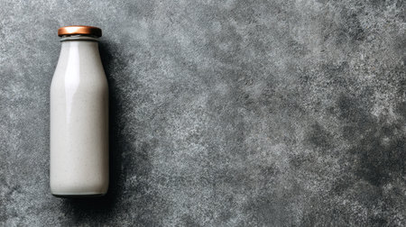 A single milk bottle sits on a mottled gray surface in an overhead composition. The bottle is opaque and white, contrasted by a brown cap. The lighting appears diffused, emphasizing the texture. This image could be used for product promotion, food-related content, or design projects. The neutral tones offer ample copy space.の素材