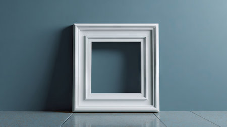 A white, square picture frame rests against a muted blue wall, creating a simple composition. The frame's clean lines and neutral color contrast the textured surface. Soft lighting suggests an interior setting. This image could be used for product mockups, artistic presentations, or editorial content.の素材