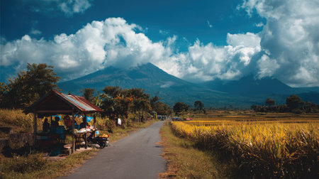 An outdoor scene features a road winding through a field towards a large mountain range under a cloudy sky. The composition highlights natural colors, with a mix of green, brown, and blue hues. The presence of a shelter suggests a recreational setting. This image may be suitable for travel, tourism, and environmental publications.の素材