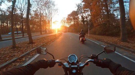 A first-person perspective captures a motorcycle ride along a road. The scene is bathed in warm sunlight from a setting sun. The composition includes a blurred background featuring trees and another motorcyclist. Suitable for editorial and commercial use, this image evokes a sense of freedom.の素材