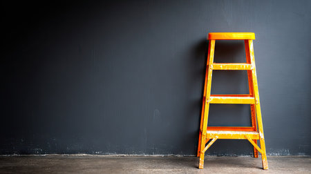 An orange step ladder stands against a dark wall in a minimalist composition. The image features a side view of the wooden structure with warm tones, contrasting with the cool background. The scene suggests a plain interior, offering potential for various commercial or editorial applications.の素材