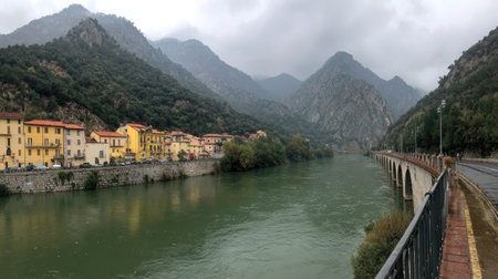 A river scene showcases a town situated along the water's edge, framed by rolling mountains. The image features a subdued color palette under an overcast sky, with the river reflecting the muted tones. It presents potential applications for environmental, travel, and editorial projects, offering a sense of tranquility.の素材