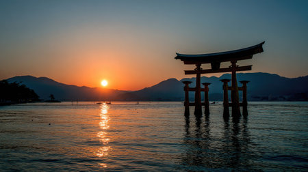 A silhouette of a traditional structure stands in calm water at sunset. The image displays warm orange and yellow hues from the setting sun, reflecting on the water's surface. Mountains are visible in the background against a blue sky. Suitable for travel articles, environmental themes, or artistic projects.の素材