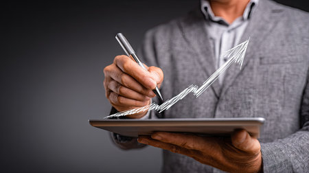 A person in a business suit is depicted using a pen to draw a rising graph on a tablet, symbolizing growth. The image uses a gray color scheme, a close-up perspective, and soft lighting to highlight the subject and the digital device. This visual is suitable for depicting business development, market analysis, or financial strategies.の素材
