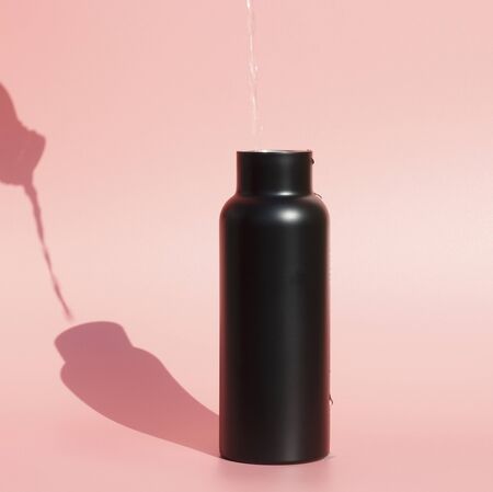 Water pours into a black reusable bottle on a pink background.の写真素材