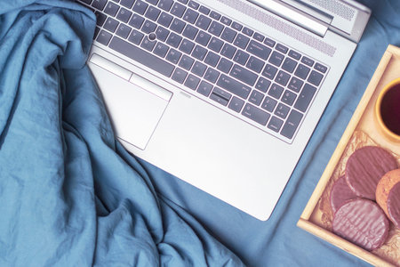 A laptop and a wooden tray with cookies and coffee are on the bed top view.