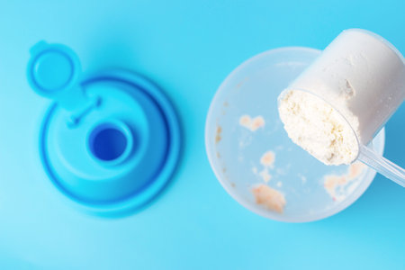 Protein powder is poured from a measuring spoon into a cocktail shaker on a blue background, top...