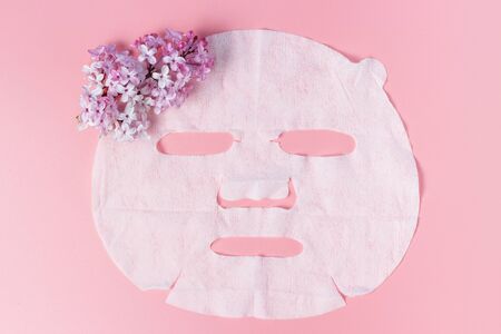 White fabric mask with a branch of lilac on a pink background close-up, skin hydration concept.の写真素材