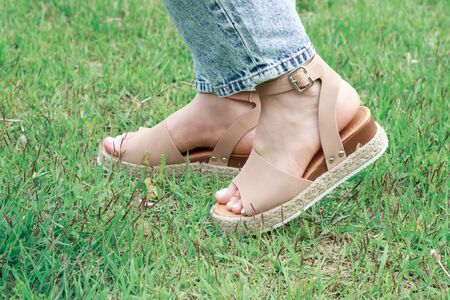 Women's feet in summer brown sandals against the background of green grass, the concept of summer shoesの写真素材