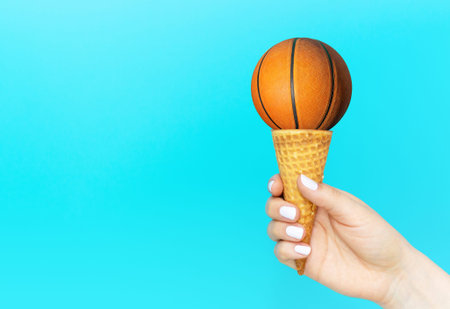 Female hand holds a ice cream cone with basketball, creative summer background.