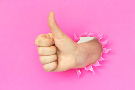 Female hand raises thumb up on a torn pink paper background.の写真素材