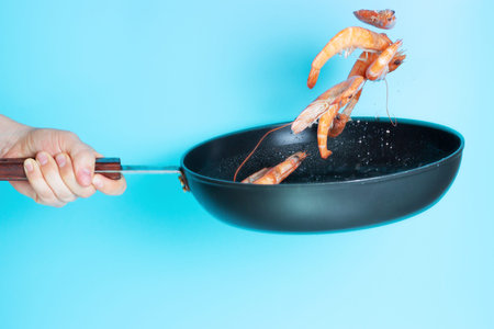 A womans hand holds frying pan and frys shrimp on blue background