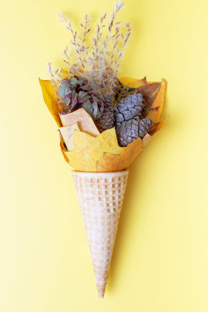 Bouquet of dry leaves and cones in waffle cone on yellow background top view.
