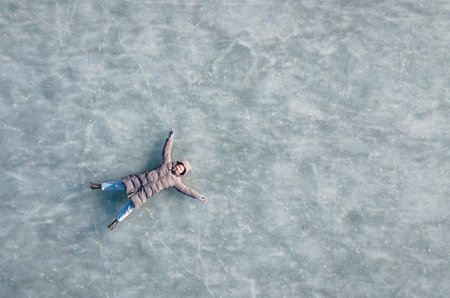 Young woman in winter down jacket lies on frozen ice, top viewの写真素材