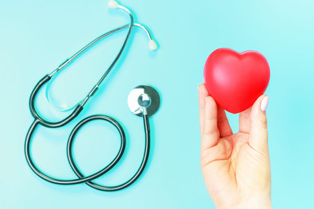 Red heart in female hands and stethoscope on blue background.