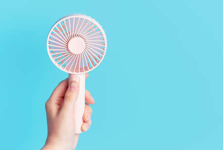 Female hand holding a pink mini fan on a blue background close-up.の写真素材