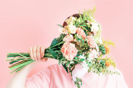 Young woman holding a bouquet of flowers covering her face on a pink background.
