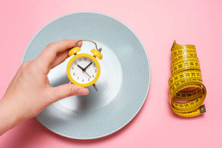 Female hand holding an alarm clock on the background of an empty plate, concept of intermittent fasting.の素材 [FY310191435137]