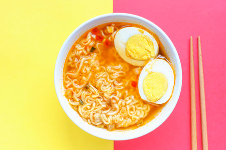 Bowl with instant noodle soup and eggs on red and yellow background close-up, top view.の写真素材
