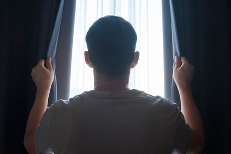Man stands at window and opens curtains rear view.の写真素材