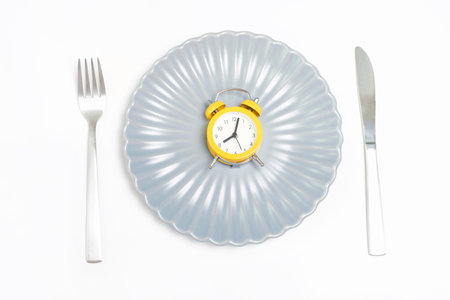 Empty plate with alarm clock, fork and knife on a white background, intermittent fasting concept.