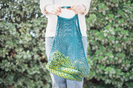 Female hand holding reusable shopping bag with vegetables on green background, zero waste concept.の写真素材