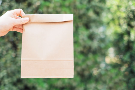 Female hand holding folded cardboard bag on a background of green leaves.の写真素材