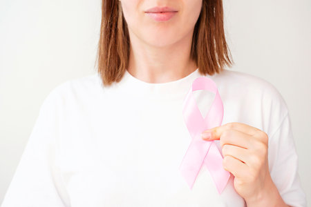 Young woman in t-shirt holding pink ribbon next to heart on white background.の写真素材