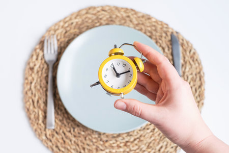 Female hand puts an alarm clock on an empty plate on white background, intermittent fasting concept.の写真素材