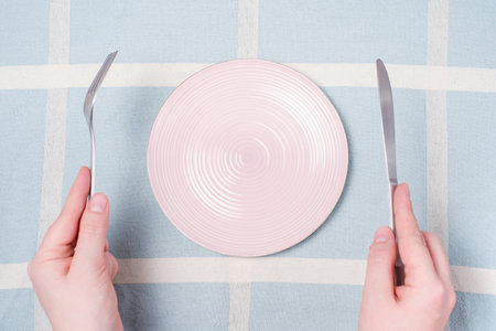 Female hands holding a fork and and knife next to an empty pink plate on a blue tablecloth background close-up top view.の写真素材