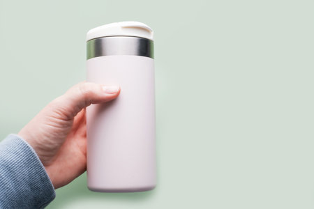 Female hand holding thermo mug or thermos on a green background side view.の写真素材