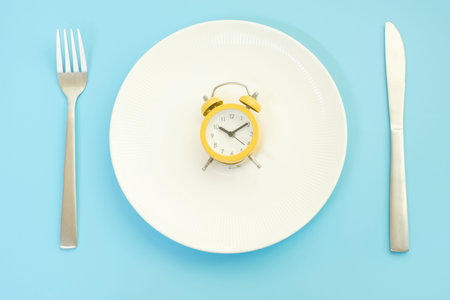Empty plate with alarm clock on blue background, intermittent fasting concept.