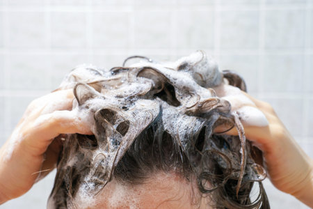A young woman washes her hair with shampoo on gray tiles background, front view.の写真素材