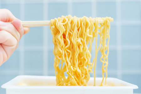 Female hand holding wooden chopsticks with instant noodles against background of blue tiles.の写真素材