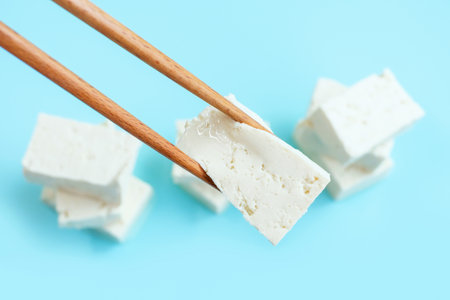 Piece of white tofu in wooden chopsticks on blue background close-up.の写真素材