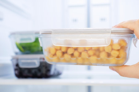 A female hand holds a glass box with chickpeas against the background of an open refrigerator, close-up.の写真素材