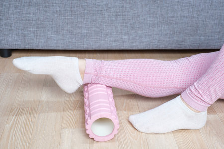 Female feet in socks rolling pink foam roller against background of gray sofa, home workout concept.