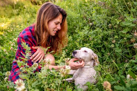 Young latin woman with her dog sitting in a flower field surrounded by tall grassの写真素材