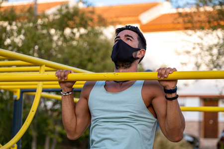Young latino man wearing pale blue tank top and black pants and mask doing exercises in a calisthenics parkの写真素材