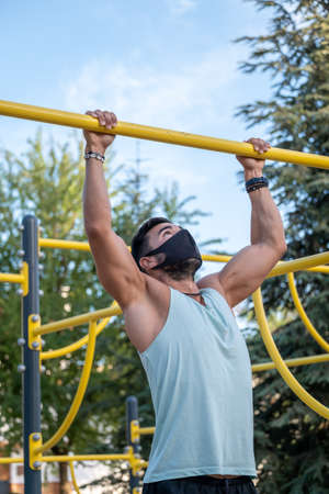 Young latino man wearing pale blue tank top and black pants and mask doing exercises in a calisthenics parkの写真素材