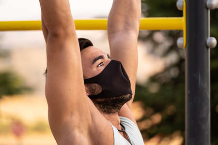 Close portrait of young latino man wearing pale blue tank top and black pants and mask doing exercises in a calisthenics parkの写真素材