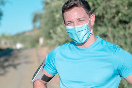 Portrait young caucasian man with mask preparing to run with smartphone in armband and a blue...