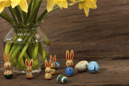 Many small Easter bunnies and easter eggs under a bouquet of daffodils against a wooden background