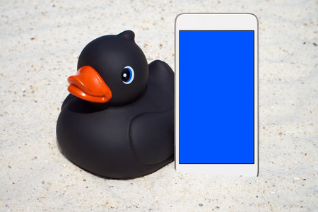 Black rubber duck and a white Smartphone with empty screen display on the beachの写真素材