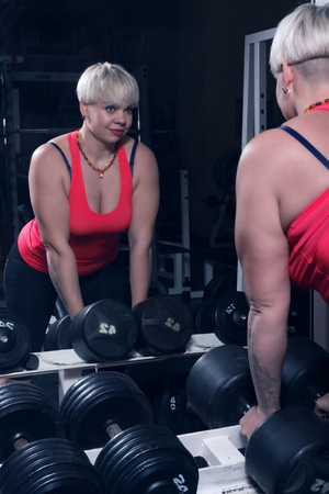 A beautiful middle-aged blonde doing exercises with dumbells and looking at her reflection at gymの写真素材