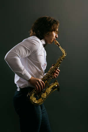 Studio portrait of young cool man wavy dark hair, in white shirt with saxophone on dark background.の写真素材